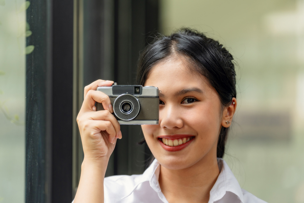 Portrait of a happy young Asian woman smiling and holding a retro vintage film camera, standing by the window with natural light, lifestyle and photography concept