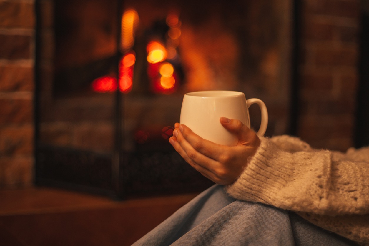 Young woman wearing white woolen knitted sweater enjoying hot tea near fireplace in a cozy living room.